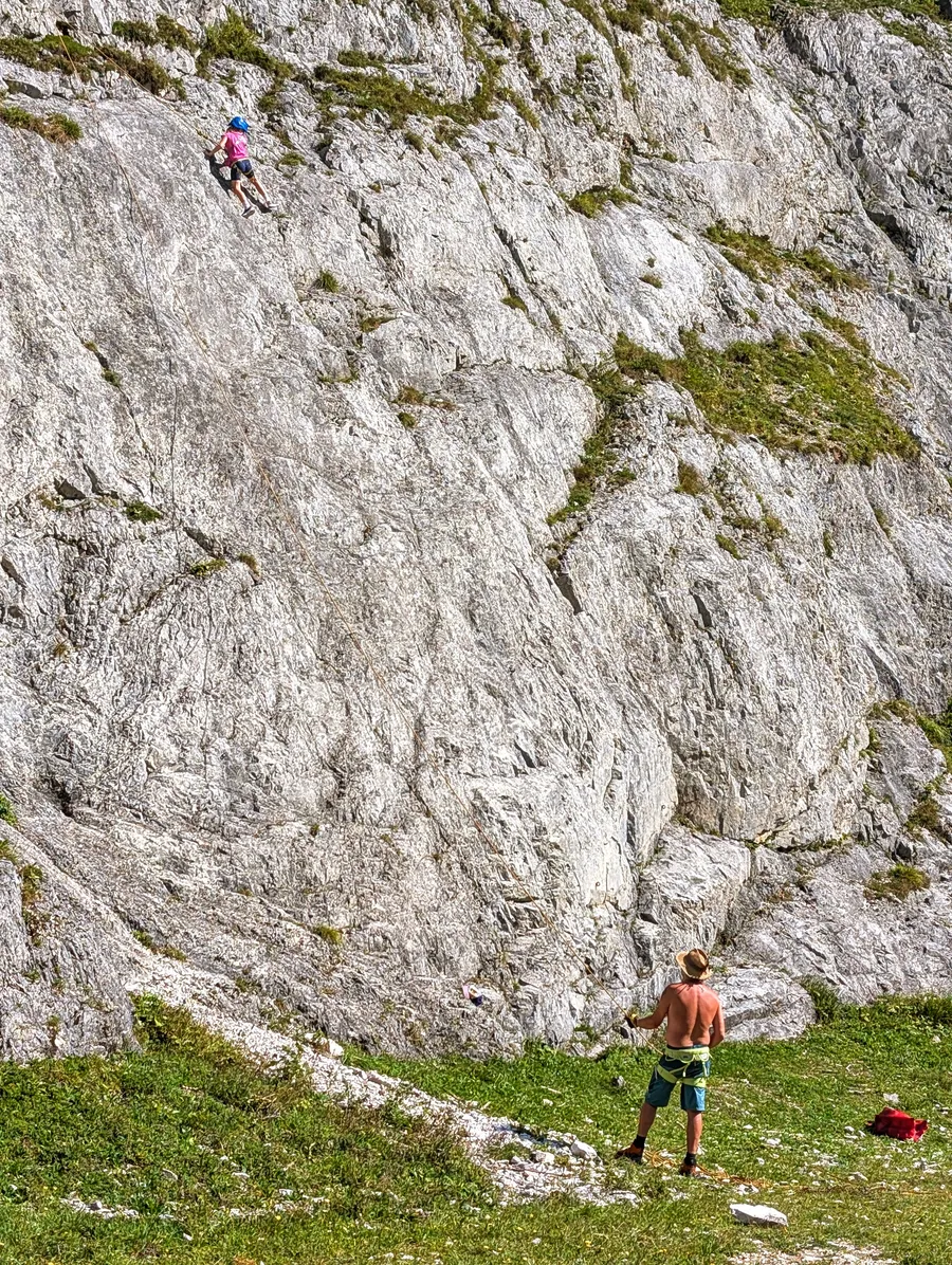 Ein Kind klettert eine Felswand hinauf, unten sichert ein Erwachsener